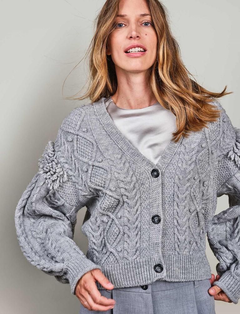 Front top half view of a woman wearing the summum balloon sleeve cardigan in the color fog/grey. This sweater has a cable knit pattern, a button down front, and fringe on the balloon sleeves.