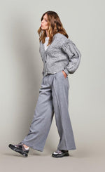 Load image into Gallery viewer, Left side full body view of a woman wearing grey pants and the summum balloon sleeve cardigan in the color fog/grey. This sweater has a cable knit pattern, a button down front, and fringe on the balloon sleeves.
