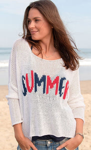 front top half view of a woman on a beach wearing the wooden ships summer loving crew top and jean shorts. This knit top is white with the word summer knit into the front center of it in red and blue. The top has a boxy silhouette and long sleeves.