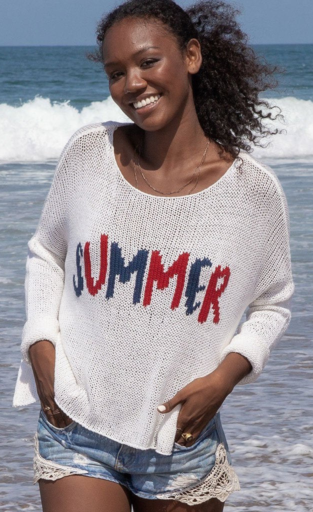 front top half view of a woman on a beach wearing the wooden ships summer loving crew top and jean shorts. This knit top is white with the word summer knit into the front center of it in red and blue. The top has a boxy silhouette and long sleeves.