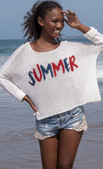 Load image into Gallery viewer, front top half view of a woman on a beach wearing the wooden ships summer loving crew top and jean shorts. This knit top is white with the word summer knit into the front center of it in red and blue. The top has a boxy silhouette and long sleeves.
