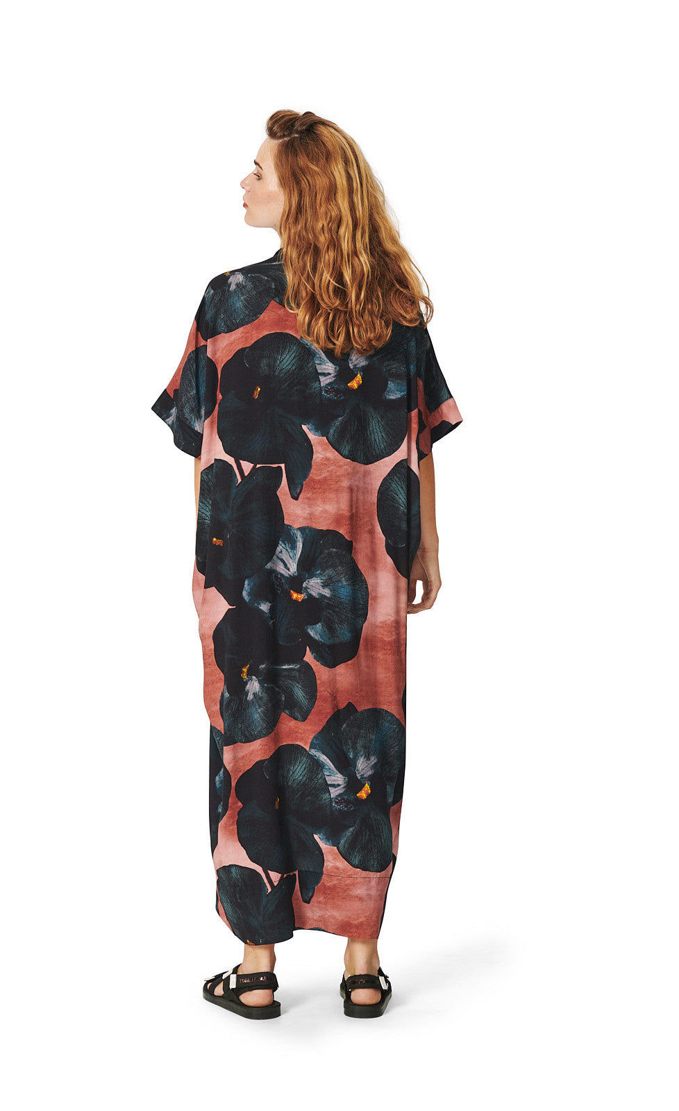 Back full body view of a woman wearing the bitte kai rand black hibiscus long dress. This dress is pink with large black hibiscus flowers all over it. The dress has elbow-length sleeves, a v-neck, and a hem that sits at the ankles.