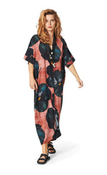 Load image into Gallery viewer, Front full body view of a woman wearing the bitte kai rand black hibiscus long dress. This dress is pink with large black hibiscus flowers all over it. The dress has elbow-length sleeves, a v-neck, and a hem that sits at the ankles.
