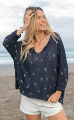 Load image into Gallery viewer, Front top half view of a woman on the beach wearing white shorts and the wooden ships mini starfish top in indigo. This top has 3/4 length sleeves, a v-neck and an oversized silhouette.
