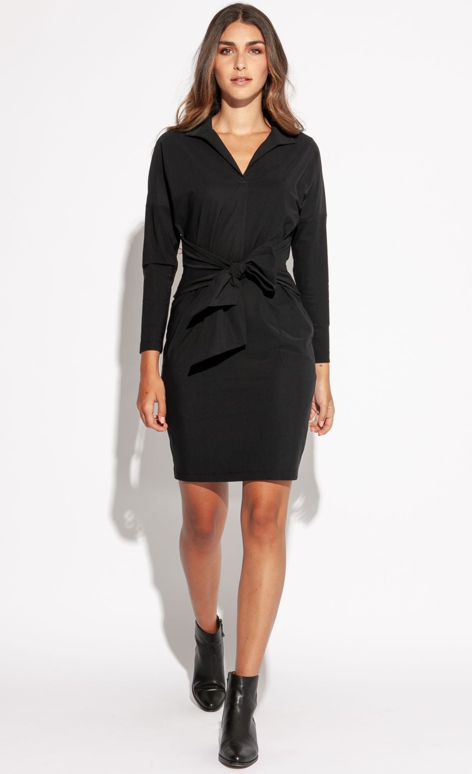 Front full body view of a woman walking forward wearing the Indies Irina Dress. This black fitted dress cuts off above the knees and has long sleeves and a large tie belt on the waist.