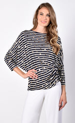 Load image into Gallery viewer, Front view of a woman with her right hand on her hip wearing white pants and the Frank Lyman Navy &amp; White Striped Knit Tunic. This striped tunic features gold dots all over it, 3/4 length dolman sleeves, and a gathering on the left side near the hip.
