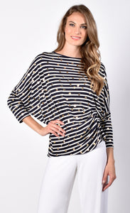 Front view of a woman with her right hand on her hip wearing white pants and the Frank Lyman Navy & White Striped Knit Tunic. This striped tunic features gold dots all over it, 3/4 length dolman sleeves, and a gathering on the left side near the hip.