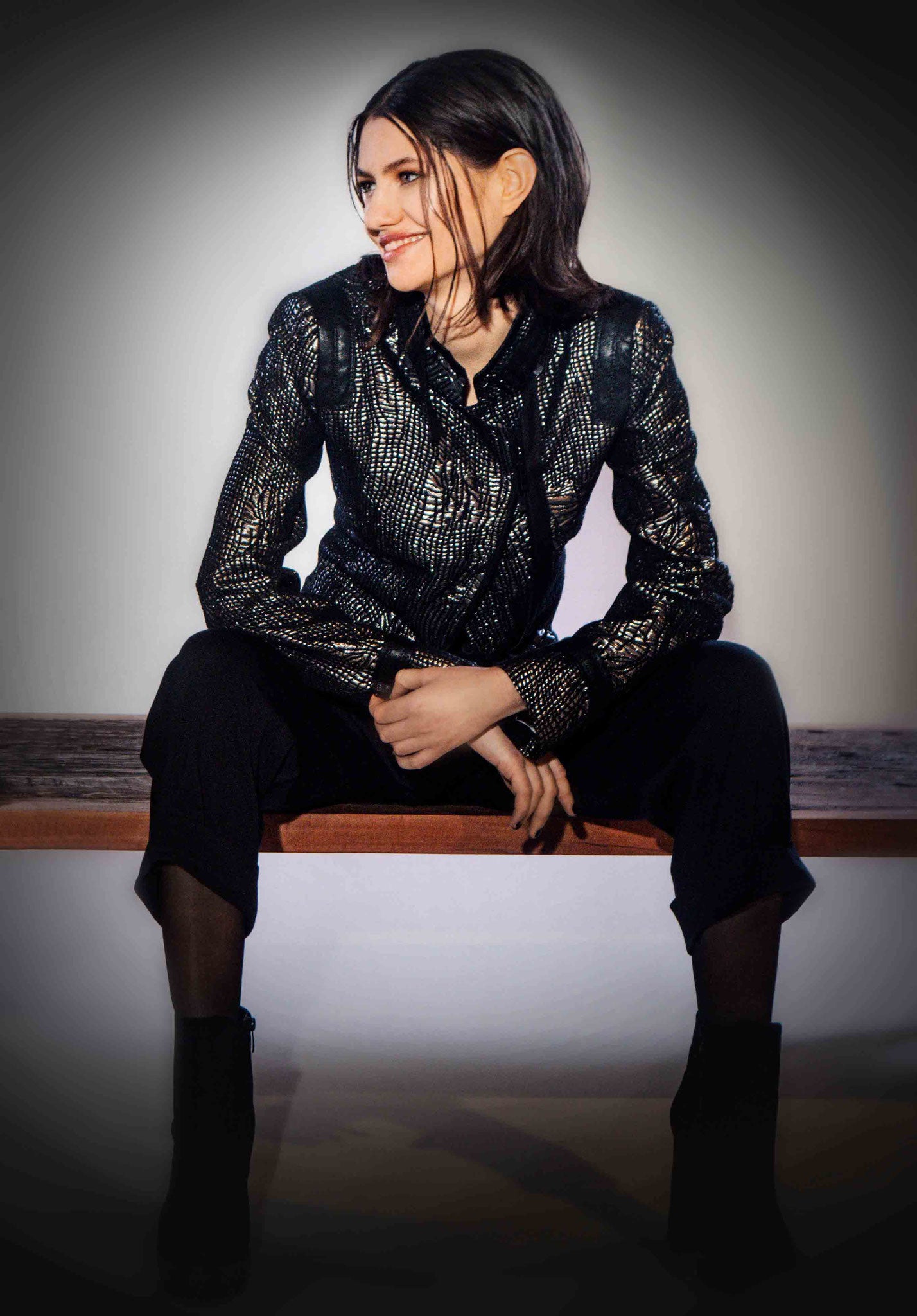 Front full body view of a woman sitting and wearing black pants and the beate heymann carbon jacket. This jacket is black with a cracked gold print all over it. The jacket is short with long sleeves, and off-center zipper, and a notched collar.