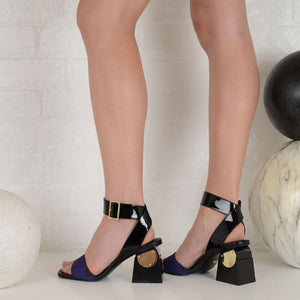 Outer and inner side view of a model wearing a pair of the kat maconie nyla sandal. This sandal has a black patent leather upper near the heel and a black patent leather ankle strap with a buckle. The upper near the square toe is blue and varnished. The heel is black with a gold circle.