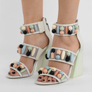 Outer and inner view of a woman wearing the kat maconie cardi high-heel. This shoe is white with three straps. Each strap is decorated with pastel colored beads. The heel is geometric with pastel green outlining.