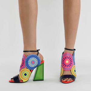 Inner and front view of a woman wearing a pair of the kat maconie frida glitch high heel. This shoe features bright mutlicolored circles on the upper with a black strap over the toes and a black ankle strap. The high heel is green.