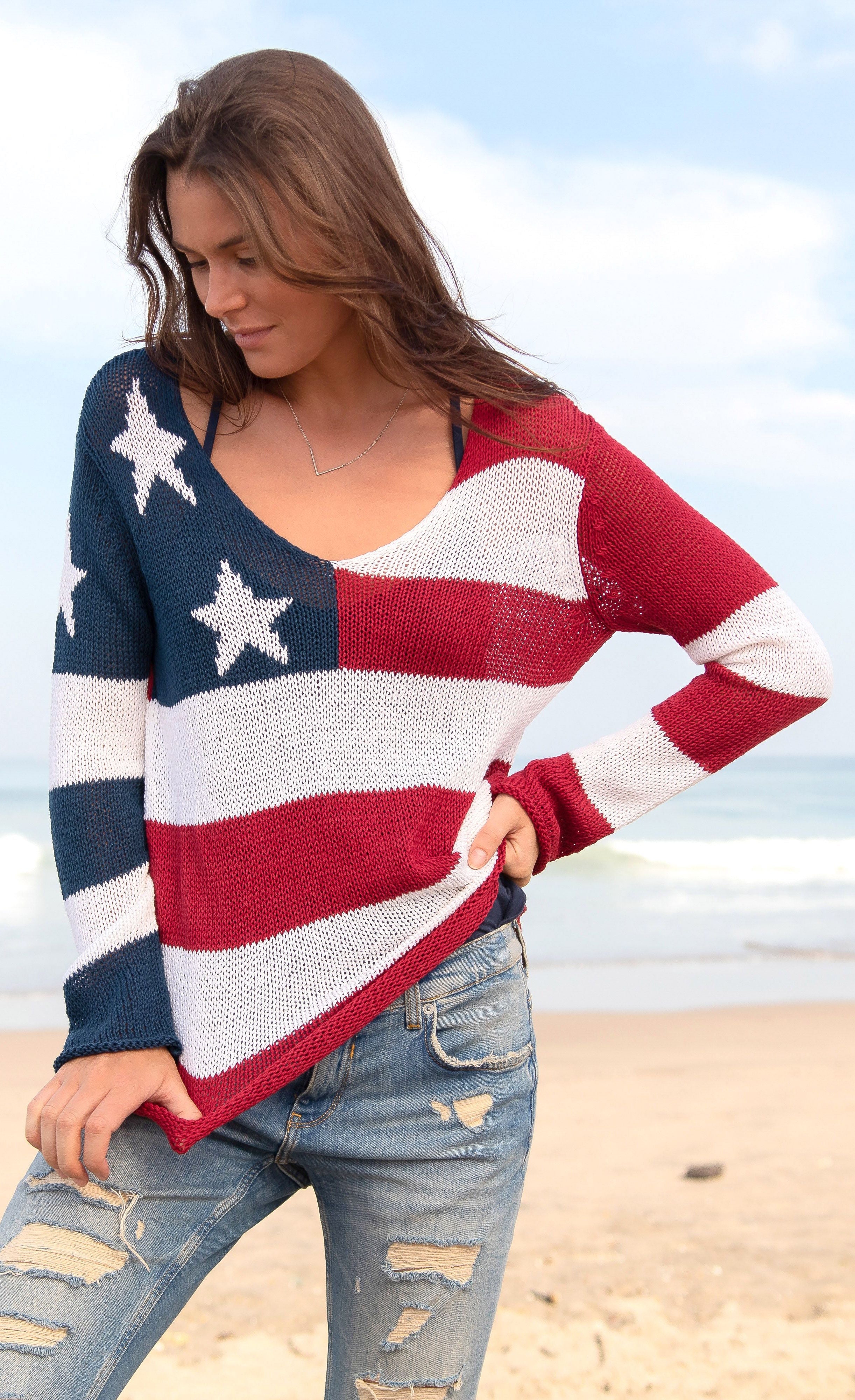 Front full body view of a woman standing on the beach wearing the wooden ships flag v cotton sweater. This sweater has the flag on it with a v-neck and long sleeves.