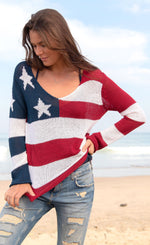 Load image into Gallery viewer, Front full body view of a woman standing on the beach wearing the wooden ships flag v cotton sweater. This sweater has the flag on it with a v-neck and long sleeves.
