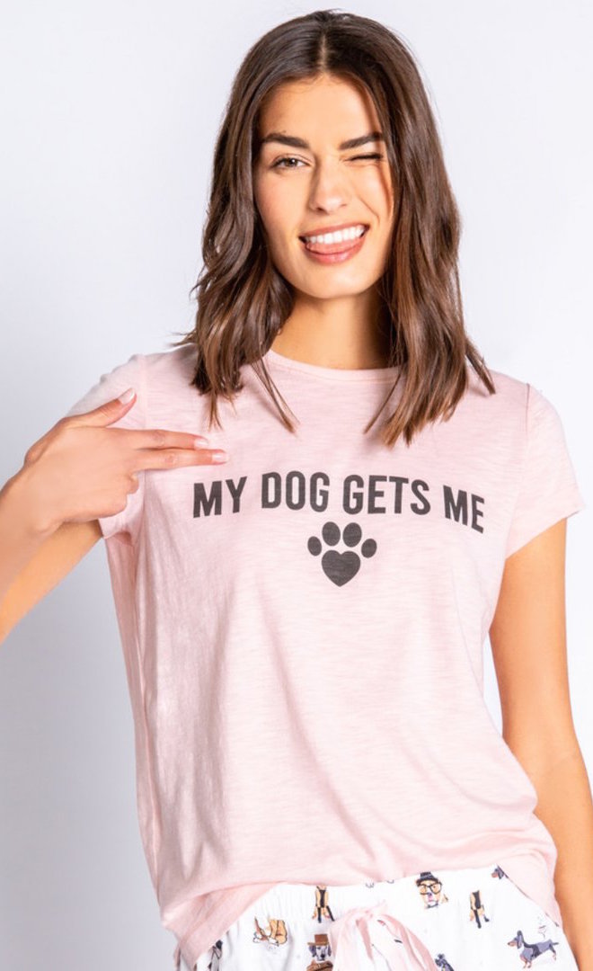 Front top half view of a woman winking and pointing to the pink tee she is wearing from PJ salvage that says My Dog Gets Me.
