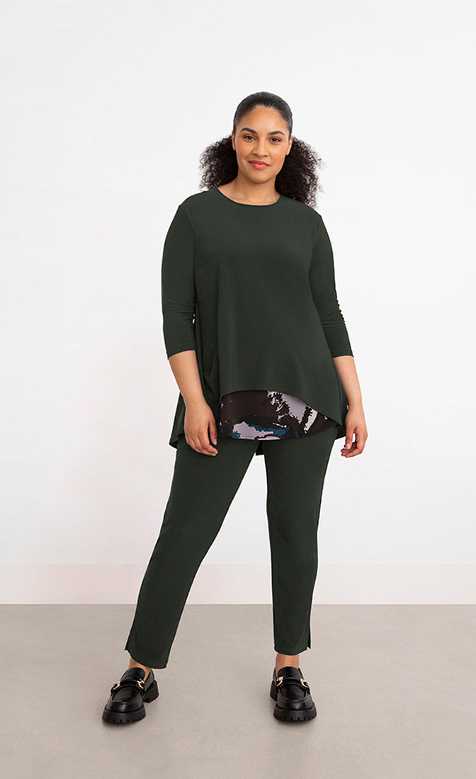 Front full body view of a woman wearing the sympli nu whisper split back top. This top is in the color green/seaweed with a mineral print peeking out from the bottom. The top has 3/4 length sleeves and a round neck.