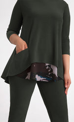 Load image into Gallery viewer, Front close up view of a woman wearing the sympli nu whisper split back top. This top is in the color green/seaweed with a mineral print peeking out from the bottom. The top has 3/4 length sleeves and a round neck. The model has her hand in the front pocket.
